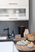 Bread and melon on wooden boards on corner of L-shaped kitchen counter with white cabinet doors and grey stone worksurface