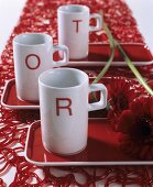Drei weiße Porzellantassen mit aufgedruckten roten Buchstaben auf roten Tabletts, Gerberas und roter Kunststoff-Tischläufer