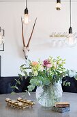Blumenstrauss in kugelförmiger Glasvase und Messing Kerzenständer mit Teelichtern auf Tisch unter Glühbirnen Pendelleuchten