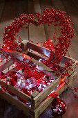 Herz-Kranz aus roten Kunstbeeren, eine Lichterkette und Christbaumkugeln in einer Holzkiste