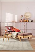 A comfortable 1950s-style lounger with cushions and throw, a side table on a patterned rug and a sideboard and a wall painted pastel purple