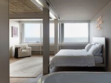 Reduziertes Schlafzimmer mit Doppelbett und weißem Polstersessel vor Panoramafenster und Meerblick