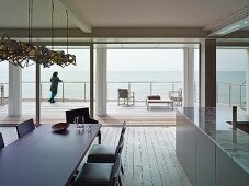 View from dining area with dark table and upholstered chairs through open sliding glass wall to wooden terrace and sea