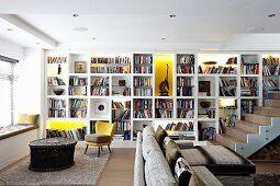 Bookcase with some compartments illuminated, sofa, armchair and ethnic table next to window in open-plan interior