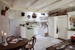 weiße Landhausküche mit Tiertrophäen an gefliesten Wänden, weiß lackierter Balkendecke und Kerzenlicht auf ovalem, antikem Tisch