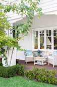 Blick vom Garten auf Veranda mit hellen Rattanmöbeln vor holzverkleidetem, weißem Landhaus