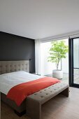 Elegant double bed with taupe headboard against black wall and taupe bedroom bench next to open sliding door with view of potted tree on patio