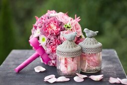 Romantischer Hochzeitsstrauß mit rosafarbenen Rosen, verstreuten Rosenblütenblättern und Glasgefäßen mit Vogelfiguren