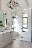Pale grey bathroom washstand below elegant mirror, chandelier and modern shower area with glass partition