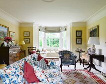 Sofa mit blumigem Stoffbezug und passende Kissen, Polstersessel und klappbarer Antiktisch in gelb getöntem Wohnzimmer mit Erker