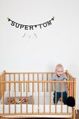 Toddler standing in cot below garland of black letters spelling name on wall