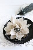 Handgenähte Seidenblume mit silbernem Knopf in Tortelettform auf Spitzenpapierdeckchen, dahinter schwarze Feder