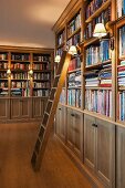 Sconce lamps with lampshades attached to floor-to-ceiling bookcases in traditional library with library ladder
