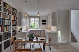 Square, rustic wooden table, vintage chairs and bookcase in front of white fitted kitchen with panelled fronts