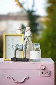 Yellow picture frame, preserving jar, bottle decorated with floral paper and horse figurine on top of pink vintage suitcase
