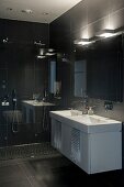 Waschtisch mit weißem Unterschrank an schwarz gefliester Wand, oberhalb Wandleuchten über Spiegel in einem Designerbad