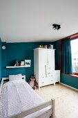 Wood-framed bed with striped bed linen and white wardrobe against petrol walls in child's bedroom