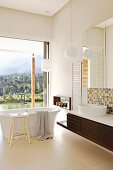 Modernes Bad mit freistehender Badewanne vor Glasschiebetür mit Panoramablick, seitlich massgefertigter Waschtisch mit Spiegelschrank