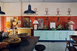 Blick über Theke mit Keramikgeschirr auf Küchen-Sideboard, dekoriert mit Tischleuchten vor rotbrauner Wand und Wandschmuck