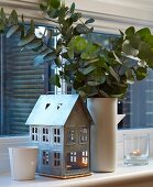 Miniaturhaus als Windlicht neben Vase mit Blätterzweigen auf Fensterbank