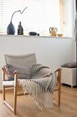 Armchair with wooden frame, linen seat and grey and white fringed blanket below window