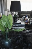 Green leaves in glass vase in front of stacked books and black table lamp on table made from pallet
