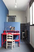 Holzstuhl und Schreibtisch im Rotton vor blauer, halbhoher Wand, dahinter Duschbereich