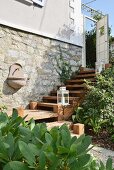 Gartenwaschbecken an Natursteinwand eines renovierten Landhauses mit Zedernholztreppe und aufgestellten weissen Laternen