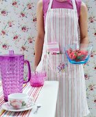 Woman wearing striped apron holding glass bowl of strawberries in front of romantic floral wallpaper
