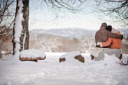 Mann und Frau sitzen Arm in Arm und blicken auf verschneite Landschaft