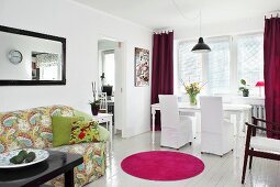 Offener Wohnbereich mit weißen Hussenstühlen und Esstisch auf weißem Dielenboden, pinkfarbener runder Teppichvorleger vor gemustertem Sofa