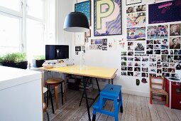 Ess- und Arbeitsplatz mit gelber Tischplatte und blauem Tritthocker vor Wand mit Fotosammlung und gerahmten Postern