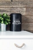Black metal tin and foliage plant on kitchen cabinet against rustic wood cladding