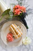 Sponge fingers tied with string and salmon-pink carnations on white plate next to bouquet of carnations on marble surface