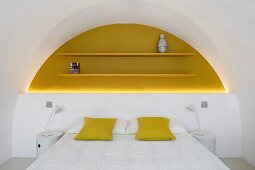 Doppelbett mit gelben Kissen vor halbhoher weißer Wand, darüber gelb lackierte Regalböden in hinterleuchteter gleichfarbiger Rundbogennische