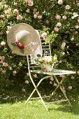 Vintage Gartenstuhl mit Blumengesteck und romantischem Strohhut vor rosafarbener Kletterrose