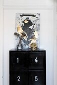 Gemälde mit Vogelmotiv und goldglänzender Totenkopf unter Glashaube auf schwarzem Schrank