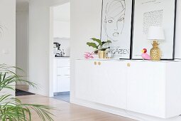 Weisser Sideboard mit Zimmerblume, Ananas-Tischlampe und angelehnten Bildern in der Diele