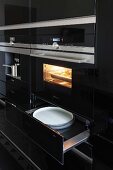 A kitchen cupboard with a shiny black surface, a built-in oven and plates in an open warming drawer