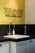 White sink in granite kitchen worksurface below decorative lettering on yellow wall