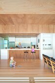 Children in open-plan interior and mother behind free-standing kitchen counter below suspended wooden ceiling, 