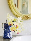 Gelbe Orchideen in blau-weißem Krug auf Waschtisch, darüber Spiegel mit geschnitztem Rahmen