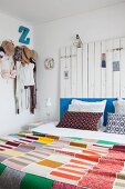 Schlafzimmer mit bunter Strick-Patchworkdecke, Betthaupt aus Holzbrettern und Kleidungsstücken an Hakenleiste