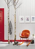 Birch trunks and stems used as a coat rack next to a an orange-coloured retro rocking chair in front of wallpaper with a shimmering fishbone pattern