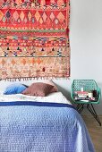 Colourful ethnic rug on wall above bed with blue bedspread and various scatter cushions