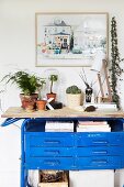 Plant arrangement and table lamp on top of blue vintage workbench below framed picture