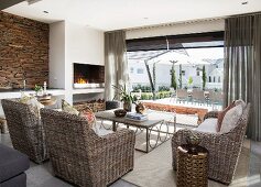 Wicker furniture, open fire and rustic stone wall in lounge with view of open-air terrace