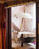 Free-standing bathtub and parasol in bathroom with patterned wallpaper and net curtains