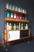 Various colourful thermos flasks, wicker bottle covers and figurines of women under glass covers on shelves above retro sideboard