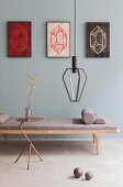 Three framed artworks on grey-blue wall above wooden couch with seat cushion, pendant lamps and metal side table